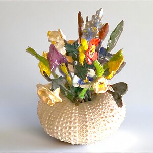 May include: A decorative arrangement of colorful, sculpted flowers in a sea urchin shell vase. The flowers include yellow, red, purple, and green elements. The vase is a light beige color and has a textured surface.