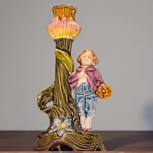 May include: A vintage ceramic candlestick holder depicting a young figure in a purple jacket and blue trousers, holding a basket. The candlestick has a floral design with a pink and yellow top, and green and brown accents. A blue flower is at the base.