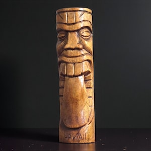 May include: A carved wooden tiki statue with a detailed face, open mouth, and protruding tongue. The word "HAWAII" is carved at the base. The statue is a light brown color and stands upright.