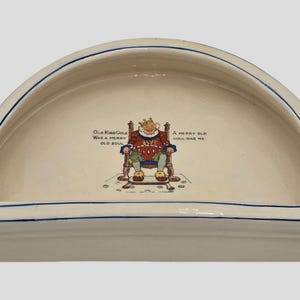 May include: A white ceramic half-round dish with a blue rim. The dish features a cartoon illustration of a king sitting on a throne with the text "Old King Cole Was a Merry Old Soul" and "A Merry Old Soul Was He".