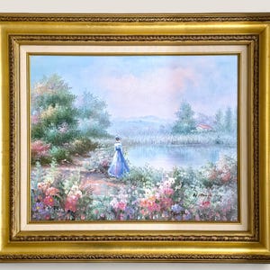 May include: A framed landscape painting featuring a woman in a blue dress near a lake. The artwork has a gold ornate frame and depicts a serene outdoor scene with colorful flowers and trees.