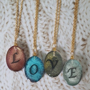 May include: Four oval-shaped pendants on thin gold-toned chains. Each pendant features a unique, textured, multi-colored resin finish with a single black letter printed on the front. Together, the pendants spell out the word LOVE in sequence.