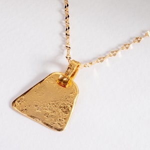 Könnte beinhalten: Eine goldene Halskette mit einem strukturierten, dreieckigen Anhänger. Der Anhänger hat eine gehämmerte Oberfläche und eine zarte Kette.