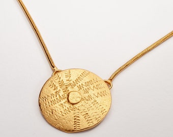 Könnte beinhalten: Eine goldene Halskette mit einem runden Anhänger mit geometrischem Muster. Der Anhänger hat eine strukturierte Oberfläche und ein kreisförmiges Muster.