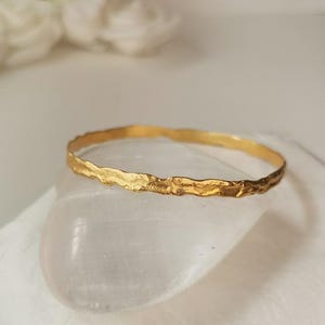 Könnte beinhalten: Ein goldfarbenes Armreif-Armband mit einem strukturierten, organischen Design. Das Armband ist kreisförmig und scheint aus Metall zu sein. Der Hintergrund ist ein weiches Weiß, das die warmen Töne des Schmucks hervorhebt. Dieses Stück eignet sich für den Alltag oder besondere Anlässe.