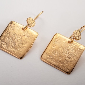 May include: A pair of gold square earrings with a textured surface and a small round accent on the top of each earring.
