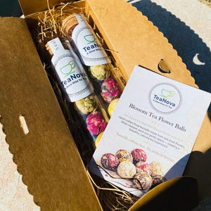 May include: A cardboard gift box containing two glass tubes of floral tea balls, with a card that reads "Blossom Tea Flower Balls". The tubes are sealed with corks and the box is filled with straw.
