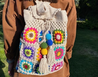 Può includere: Uno zaino bianco all'uncinetto con toppe colorate a quadrati della nonna e un fiocco bianco. Lo zaino ha una chiusura a coulisse ed è decorato con pompon colorati.