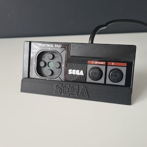 May include: Black Sega Genesis controller with a grey control pad and the text "CONTROL PAD" and "SEGA" printed on it.