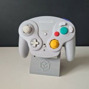 May include: A white Nintendo GameCube controller with a yellow, red, blue, and green button layout. The controller is on a grey stand with the GameCube logo. The controller has the text "WAVEBIRD" printed on the bottom.