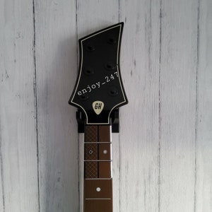 May include: Black electric guitar headstock with a brown fretboard and white buttons. The headstock has the text "enjoy_247" and a white logo with the letters "GH".