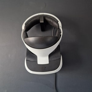 May include: A white and black VR headset on a charging dock. The headset has a curved design with a black interior and a white exterior. The charging station is black and white with the "SONY" logo.