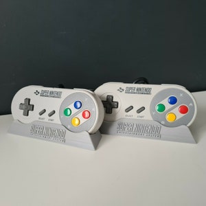 May include: Two grey Super Nintendo Entertainment System controllers with grey and white buttons, including a green, red, and yellow button, are displayed on a grey stand.