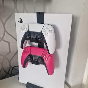 May include: Two PlayStation 5 DualSense controllers mounted on a white wall mount. The top controller is white and the bottom controller is pink.