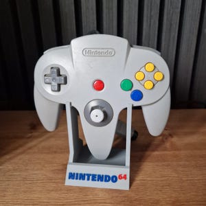 May include: A grey Nintendo 64 controller with a stand, featuring the text "NINTENDO 64" in blue. The controller has a directional pad, joystick, and coloured buttons. The stand is designed to hold the controller upright.