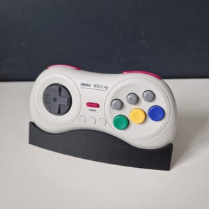 May include: A white wireless Sega Genesis controller with a black and grey d-pad, six buttons, and a red start button. The controller is labelled "8BitDo M30 2.4g" and is sitting on a black stand.