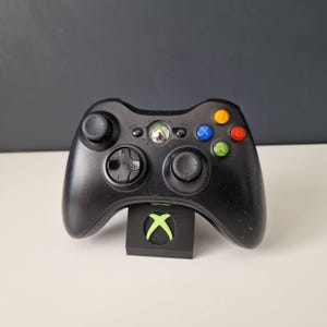 May include: A black Xbox 360 controller with green, blue, red and orange buttons. The controller is sitting on a black stand with a green Xbox logo.