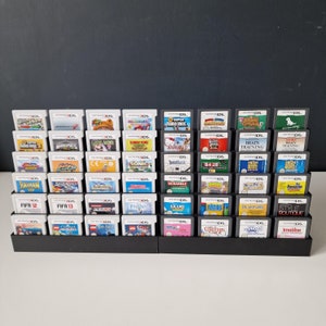 May include: A black plastic storage shelf with 42 Nintendo DS and 3DS game cartridges. The cartridges are organised in rows and columns. The shelf has a black background and is made of plastic.