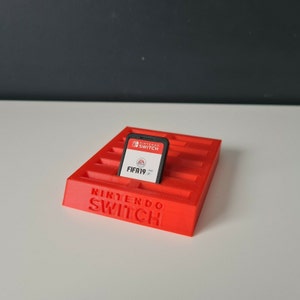 May include: A red plastic game cartridge holder with the words "Nintendo Switch" printed on the front. A black Nintendo Switch game cartridge is in the holder. The game cartridge is for FIFA 19.