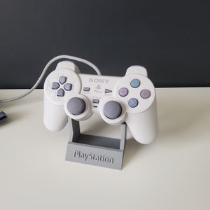 May include: A white PlayStation controller with grey buttons and a grey PlayStation logo on the stand. The controller is on a grey stand with the word "PlayStation" printed on it.