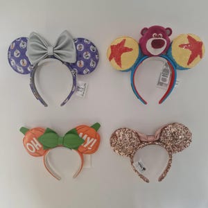 May include: Four Disney-themed ear headbands. One has Dalmatian print ears and a silver bow. Another has a plush bear and yellow ears with red stars. A third has orange pumpkin ears with a green bow and the text "OH MY!". The last has rose gold sequin ears and a pink bow.