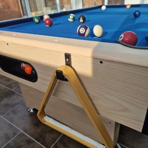 May include: A wooden pool table with a blue felt surface and a wooden triangle rack hanging on the side. The table has a black felt lining around the edge and a wooden frame. There are several billiard balls on the table, including a ball with the number 12 on it.