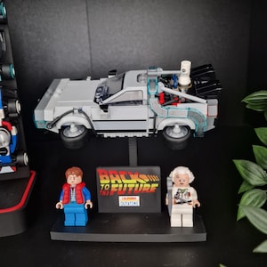 May include: A Lego model of the DeLorean time machine from the film "Back to the Future," with two Lego figures and a sign that reads "Back to the Future California Outatime." The car is silver with black and clear accents.
