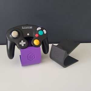 May include: A black Nintendo GameCube controller with a purple stand. The controller has a white button and a yellow button. The stand has a cube logo.
