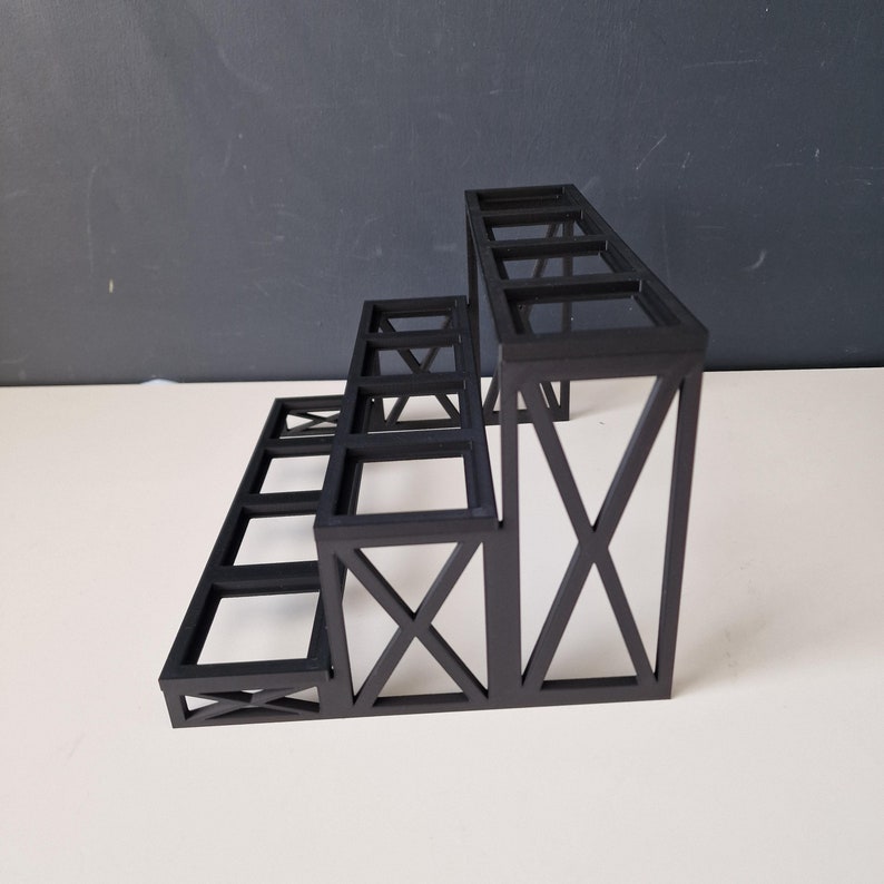 Puede incluir: Estanter&iacute;a de metal negro de tres niveles con un dise&ntilde;o geom&eacute;trico. La estanter&iacute;a est&aacute; hecha de finas barras de metal y tiene una forma rectangular.