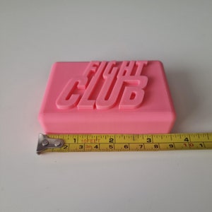 May include: A pink rectangular soap bar with the words "Fight Club" embossed on the top. The soap bar is sitting on a white surface next to a yellow measuring tape.