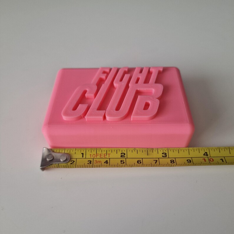 May include: A pink rectangular soap bar with the words "Fight Club" embossed on the top. The soap bar is sitting on a white surface next to a yellow measuring tape.