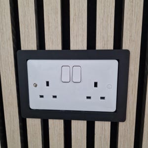 May include: A white double electrical socket with two switches, set in a black frame. The socket is mounted on vertical wood paneling with alternating light and dark wood strips.