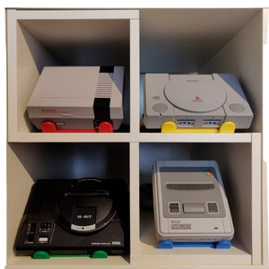 May include: Four retro video game consoles on white shelves. The top shelf has a grey Nintendo Entertainment System and a grey Playstation console. The bottom shelf has a black Sega Genesis and a grey Super Nintendo Entertainment System.