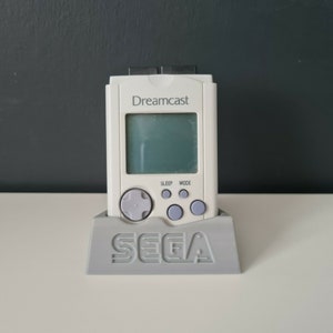 May include: A white Sega Dreamcast handheld console with a grey stand. The console has a small screen, a directional pad, and buttons labelled "Sleep" and "Mode".