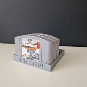 May include: A grey plastic Nintendo 64 game cartridge holder with four slots. The holder has the words "NINTENDO 64" printed on the front. The holder is filled with four grey game cartridges. The cartridge on the top has a label that reads "F1 WORLD GRAND PRIX".