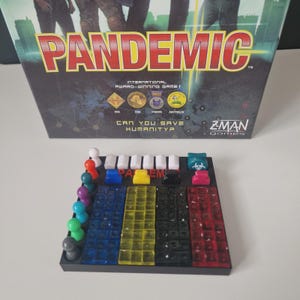 May include: A board game box titled "PANDEMIC" with red and yellow lettering. The game board shows colored squares and game pieces. The box also includes the text "CAN YOU SAVE HUMANITY?"