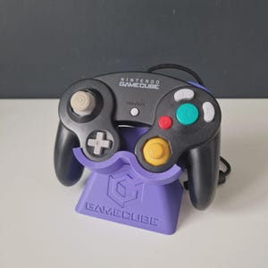 May include: A black Nintendo GameCube controller with a purple stand. The controller features a white analogue stick, a yellow button, and a directional pad. The stand displays the GameCube logo and text.