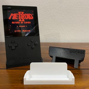 May include: A black handheld gaming console with a screen displaying the game "Metroid II: Return of Samus". The console is sitting on a white stand and a black stand with the word "pocket" printed on it.