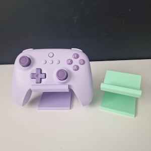 May include: A lilac video game controller with purple buttons and a cross-shaped directional pad. The controller sits on a lilac stand, with a separate mint green stand to the right.