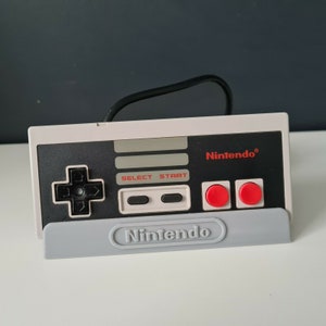 May include: A white and black Nintendo NES controller with a grey stand. The controller has a black directional pad, two black buttons labelled "Select" and "Start", and two red buttons. The stand has the word "Nintendo" printed on it.