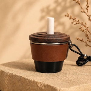 Op de afbeelding: Een bruin en zwart bekerachtig accessoire met een houten bovenkant en een wit cilindrisch stuk dat omhoog steekt. De beker heeft een bruine lederachtige band en een zwarte basis. Een zwart-wit koord is aan de zijkant bevestigd.