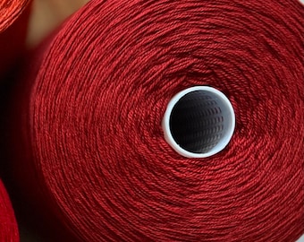 May include: A spool of red yarn wound tightly around a white cardboard tube. The yarn is smooth and has a subtle sheen.
