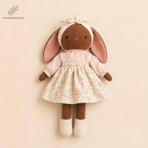 May include: A brown fabric doll with long ears, wearing a matching headband, a long-sleeved blouse, and a floral print pinafore dress in shades of pink and cream. The doll has embroidered facial features and light-coloured fabric shoes.