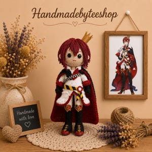 May include: Crocheted doll of a character with red hair, a crown, and a red cape. The doll is wearing a white and black outfit with gold accents. The image includes a framed picture of the character, a small chalkboard sign, and the text "Handmadebyteeshop".