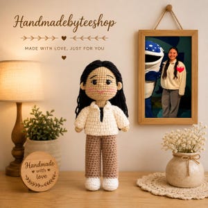 Puede incluir: Muñeca de ganchillo hecha a mano con cabello negro, que lleva una camisa color crema y pantalones marrones. La muñeca está de pie frente a una pared con una foto enmarcada y un letrero de madera que dice "Handmade with love". El texto "Handmadebyteeshop" está en la parte superior.