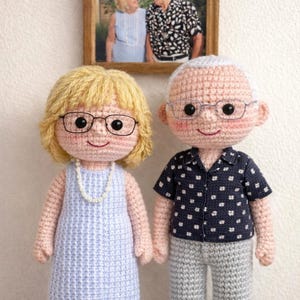 Puede incluir: Muñecos de ganchillo hechos a mano de una pareja de ancianos, cada uno con gafas. La muñeca de la mujer tiene el pelo rubio, un vestido azul y un collar de perlas. El muñeco del hombre lleva una camisa azul marino con lunares blancos y pantalones grises. Una foto enmarcada cuelga en el fondo.