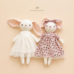 May include: Two plush rabbit dolls with embroidered facial features. The left doll wears a white lace dress. The right doll wears a pink floral dress with a matching headband. Both have long, soft ears and pink cheeks. Text reads HandmadeByTeeShop.