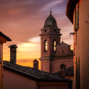 Può includere: Una vista di un piccolo paese in Italia al tramonto. L'immagine mostra una chiesa con un campanile e una cupola. La chiesa è sullo sfondo, e in primo piano si vede una strada stretta con edifici su entrambi i lati. Il cielo è di un arancione e rosa vibrante.