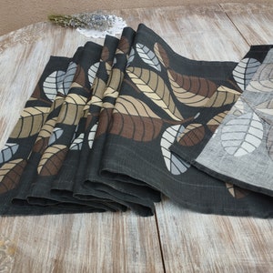 Op de afbeelding: Een zwarte linnen tafelloper met een bladmotief in tinten bruin, beige en grijs. De loper is gevouwen en wordt getoond op een houten oppervlak.