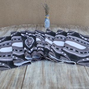 May include: A set of folded fabric placemats featuring a repeating abstract pattern in shades of black, grey, and white. The design consists of curved lines, solid shapes, and small circular dots arranged in a flowing, layered composition.